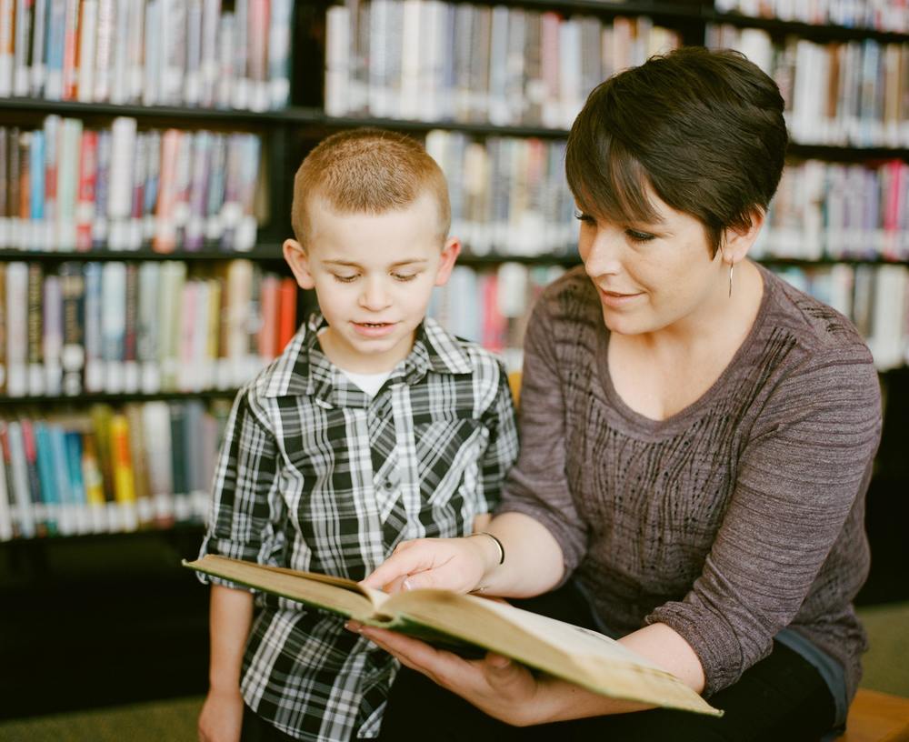 Mother reading to her son as he listens intently. Books in the background are slightly blurred creating the perfect atmosphere to instill a lifelong love of reading. The 3 - 6 year old boy is engaged, happy, the moment is captured in this photo creating a lifelong memory.